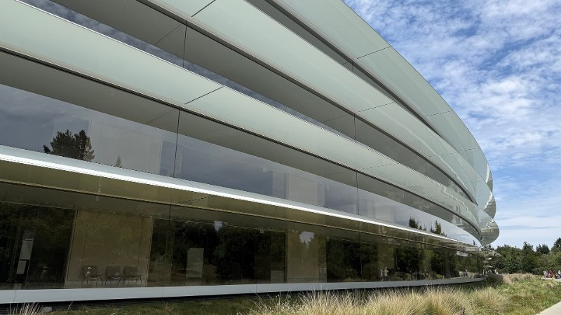 La façade vitrée incurvée du bâtiment circulaire d’Apple Park, avec des graminées au premier plan et un ciel nuageux