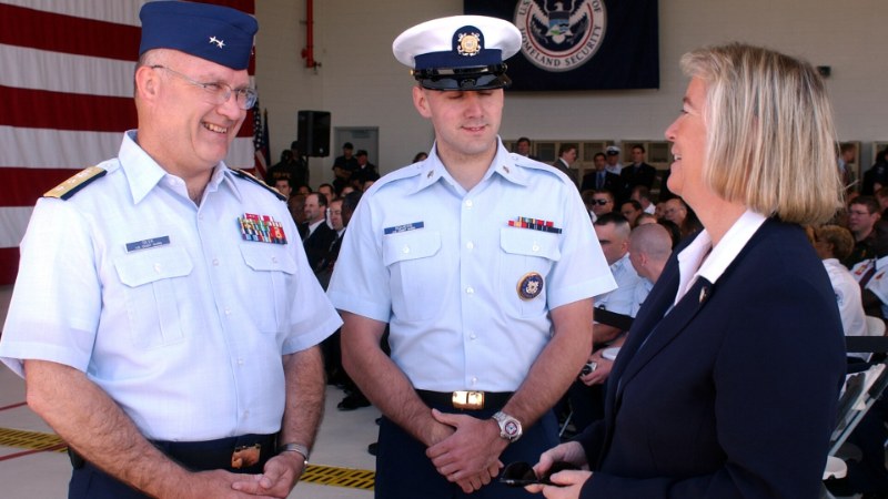 Deux personnes en uniforme et une femme en tailleur, devant une bannière au sceau du département de la Sécurité intérieure des États-Unis