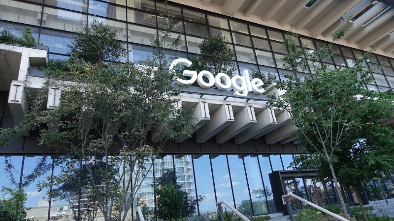 Enseigne « Google » en lettres blanches sur la façade vitrée d’un immeuble de bureaux, derrière des arbres plantés sur des terrasses en béton, avec des marches et un garde-corps au premier plan