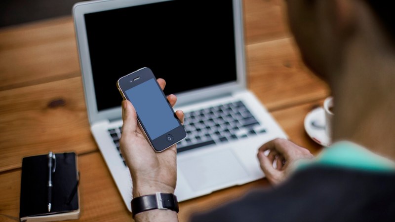 Personne vue de dos tenant un smartphone devant un ordinateur portable ouvert sur une table en bois