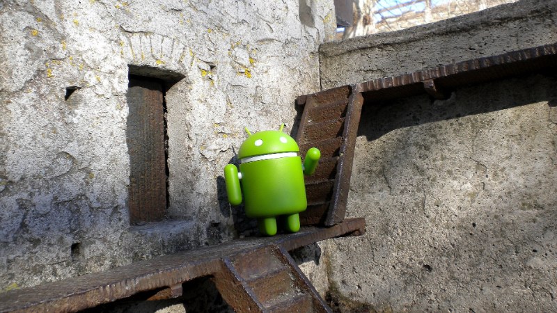 Petite figurine verte du robot Android sur une structure métallique, devant un mur de pierre
