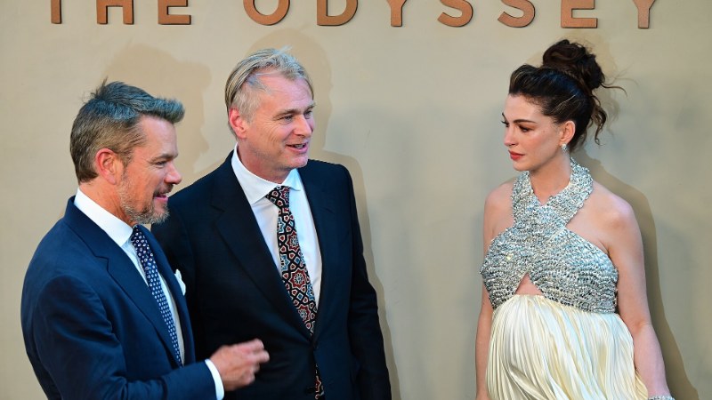 Matt Damon, Christopher Nolan et Anne Hathaway debout devant une enseigne géante portant le titre « THE ODYSSEY », sur un mur beige ; les trois posent côte à côte, Nolan au centre en costume sombre et cravate à motifs