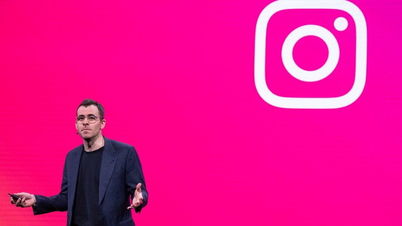 Adam Mosseri debout sur une scène devant un fond rose vif, veste sombre et lunettes, un bras tendu ; à droite, un grand logo Instagram blanc.