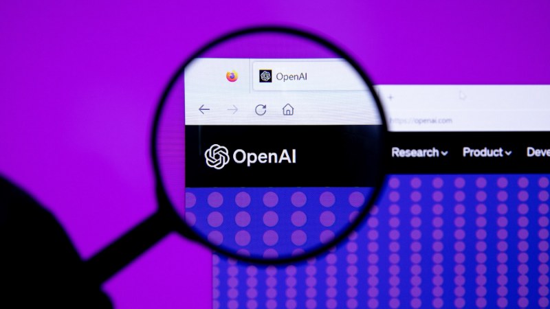 Le logotype OpenAI affiché sur un écran d’ordinateur, grossi par une loupe tenue à la main, sur un fond violet uni