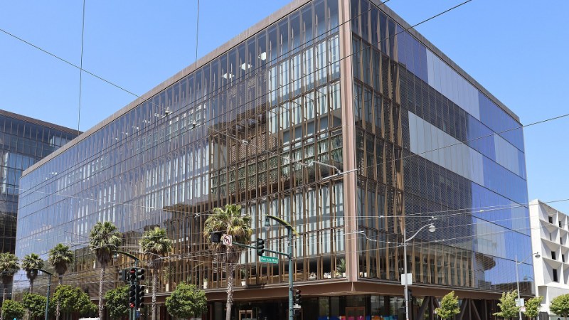 Immeuble de bureaux vitré aux montants de bronze, à l'angle de deux rues de San Francisco, sous un ciel bleu ; des palmiers et un feu de signalisation au premier plan, quelques piétons sur le trottoir