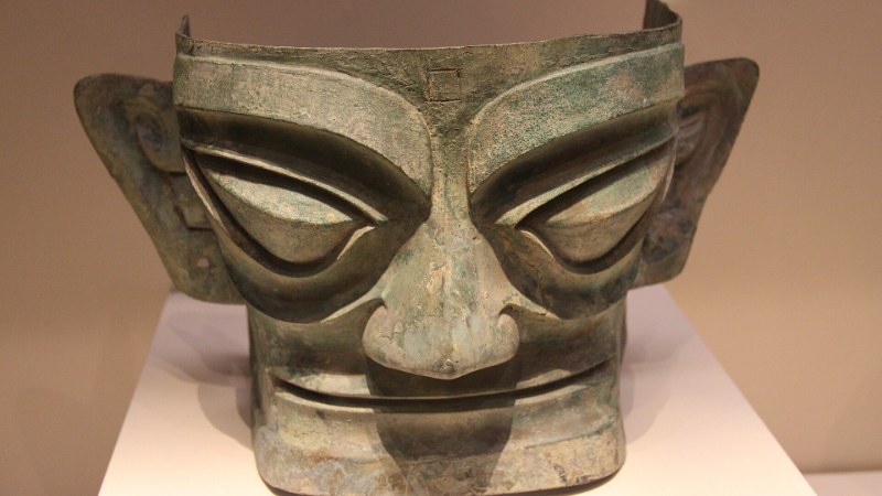 Masque de bronze de Sanxingdui vu de face : grands yeux en amande, oreilles déployées et bouche fendue, photographié sur un socle blanc de musée