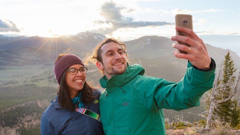 Deux randonneurs souriants prennent un selfie au smartphone devant des montagnes au coucher du soleil